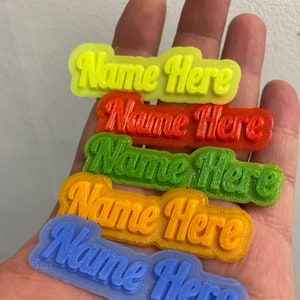 May include: Five plastic name tags in different colours, yellow, orange, green, red, and blue. Each tag says "Name Here" in a bold, cursive font.