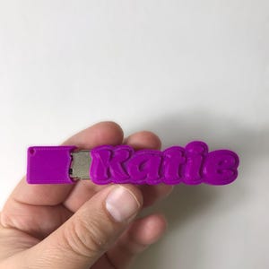 以下が含まれることがあります： 「Katie」という名前がバブルフォントで書かれた紫色のUSBドライブ。USBドライブは白い背景を背景に手で持たれています。USBドライブは無地の紫色です。