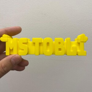May include: A bright yellow, three-dimensional plastic sign featuring the text "MS TOBLI" in bold, block capital letters. The sign has a small, cloud-like decorative shape attached to both the far left and far right ends of the lettering.