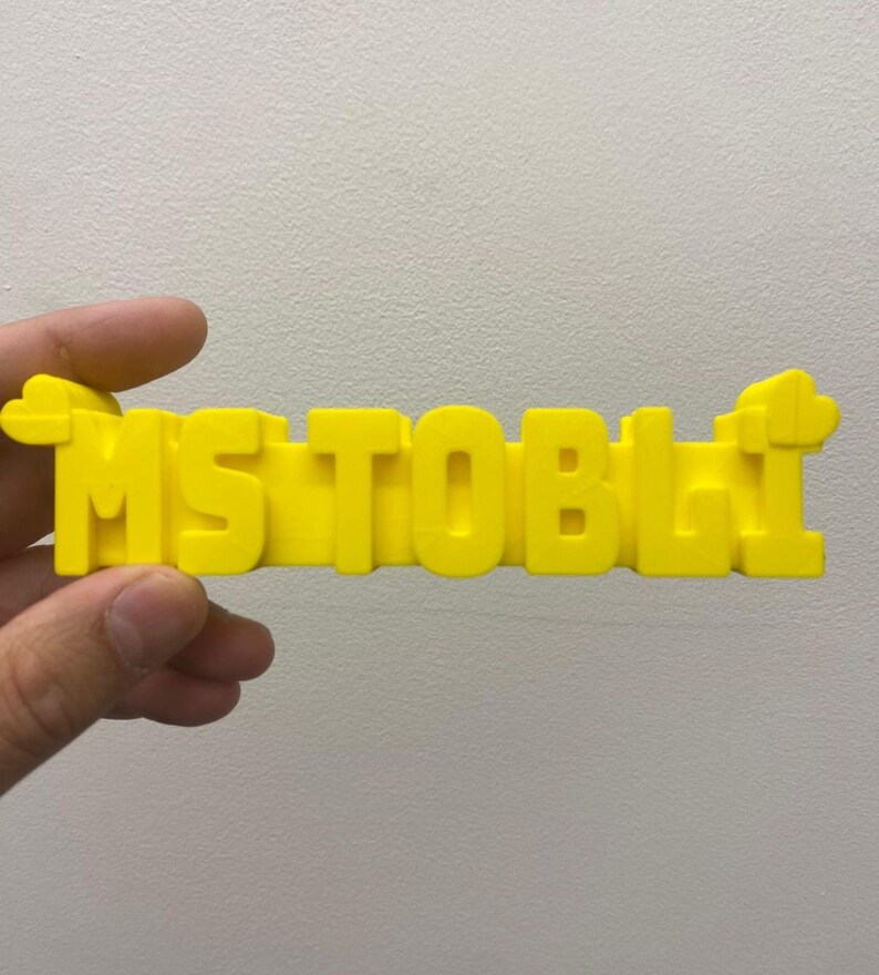 May include: A bright yellow, three-dimensional plastic sign featuring the text "MS TOBLI" in bold, block capital letters. The sign has a small, cloud-like decorative shape attached to both the far left and far right ends of the lettering.