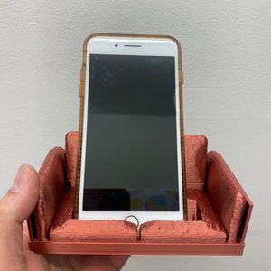 Puede incluir: Un soporte para teléfono impreso en 3D con forma de sofá, con un teléfono inteligente blanco descansando en el soporte. El soporte es de color marrón rojizo.