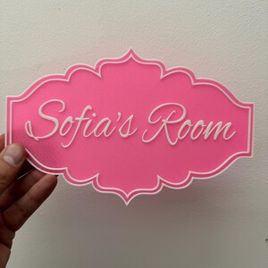 以下が含まれることがあります： 白い装飾的な縁取りと「Sofia's Room」という白い筆記体で書かれた文字が書かれたピンクのドアサイン