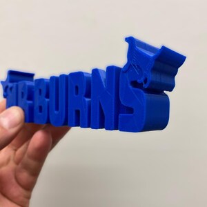May include: A three-dimensional blue plastic sign featuring the text MR BURNS in a bold, blocky font. The letter S on the right side is integrated with a stylized graphic of a shopping cart, creating a cohesive, solid blue object.