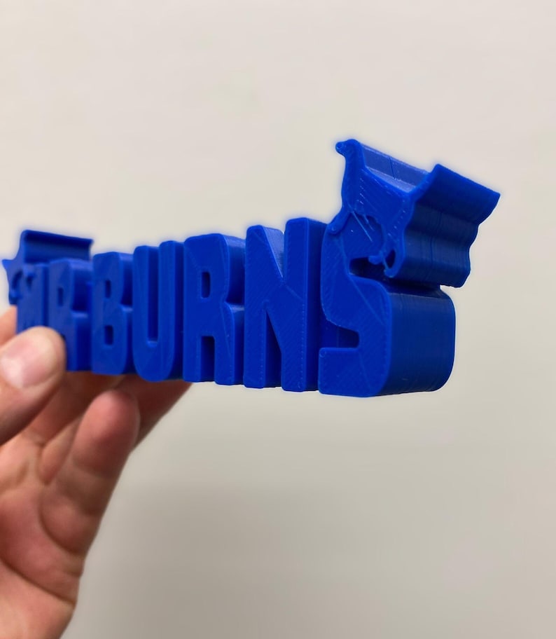 May include: A three-dimensional blue plastic sign featuring the text MR BURNS in a bold, blocky font. The letter S on the right side is integrated with a stylized graphic of a shopping cart, creating a cohesive, solid blue object.
