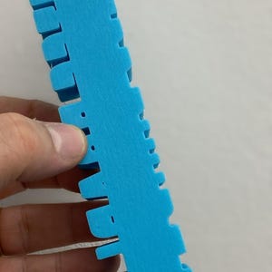 May include: A bright blue, 3D-printed plastic object with a long, rectangular base and a series of evenly spaced, protruding vertical tabs along one side. The surface has a visible, slightly textured, layered finish characteristic of additive manufacturing.