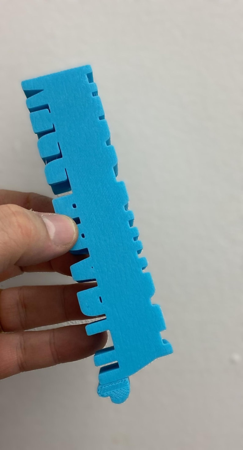 May include: A bright blue, 3D-printed plastic object with a long, rectangular base and a series of evenly spaced, protruding vertical tabs along one side. The surface has a visible, slightly textured, layered finish characteristic of additive manufacturing.