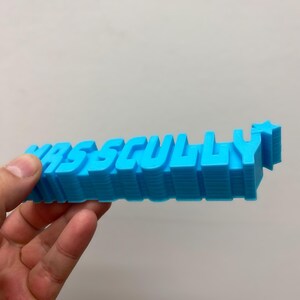 May include: A bright blue 3D printed sign featuring the text "MRSS CULLY" in a bold, blocky font. The letters are extruded to create a thick, layered appearance, and a small five-pointed star is attached to the end of the final letter Y.