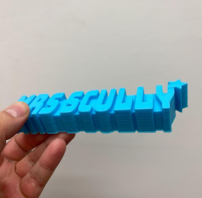 May include: A bright blue 3D printed sign featuring the text "MRSS CULLY" in a bold, blocky font. The letters are extruded to create a thick, layered appearance, and a small five-pointed star is attached to the end of the final letter Y.