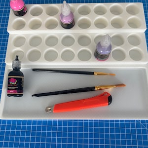 Könnte beinhalten: Ein weißer Plastik-Organizer mit mehreren Fächern zum Aufbewahren von kleinen Flaschen Farbe. Der Organizer hat zwei Ebenen und steht auf einer blauen Schneidematte. Es gibt drei Flaschen Farbe, eine rosa, eine lila und eine schwarze. Außerdem gibt es zwei Pinsel und ein Cuttermesser.