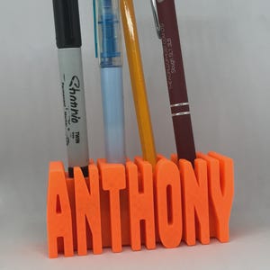 Puede incluir: Un organizador de escritorio naranja impreso en 3D con el nombre "ANTHONY" en letras gruesas. Contiene un rotulador negro, un bolígrafo azul, un lápiz amarillo y un bolígrafo burdeos.