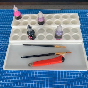 May include: A white plastic paint palette with circular wells, holding small bottles of ink in pink, purple, and black. Two paint brushes and an orange utility knife are also present.