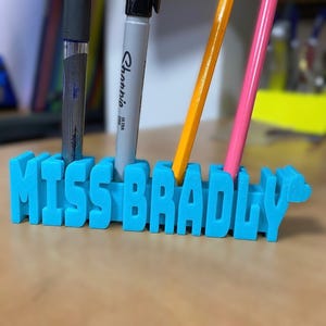 May include: A bright blue 3D printed desk organizer featuring the name MISS BRADLY in raised block letters. The organizer has four vertical slots holding a black pen, a gray marker, an orange pencil, and a pink pencil in an upright position.