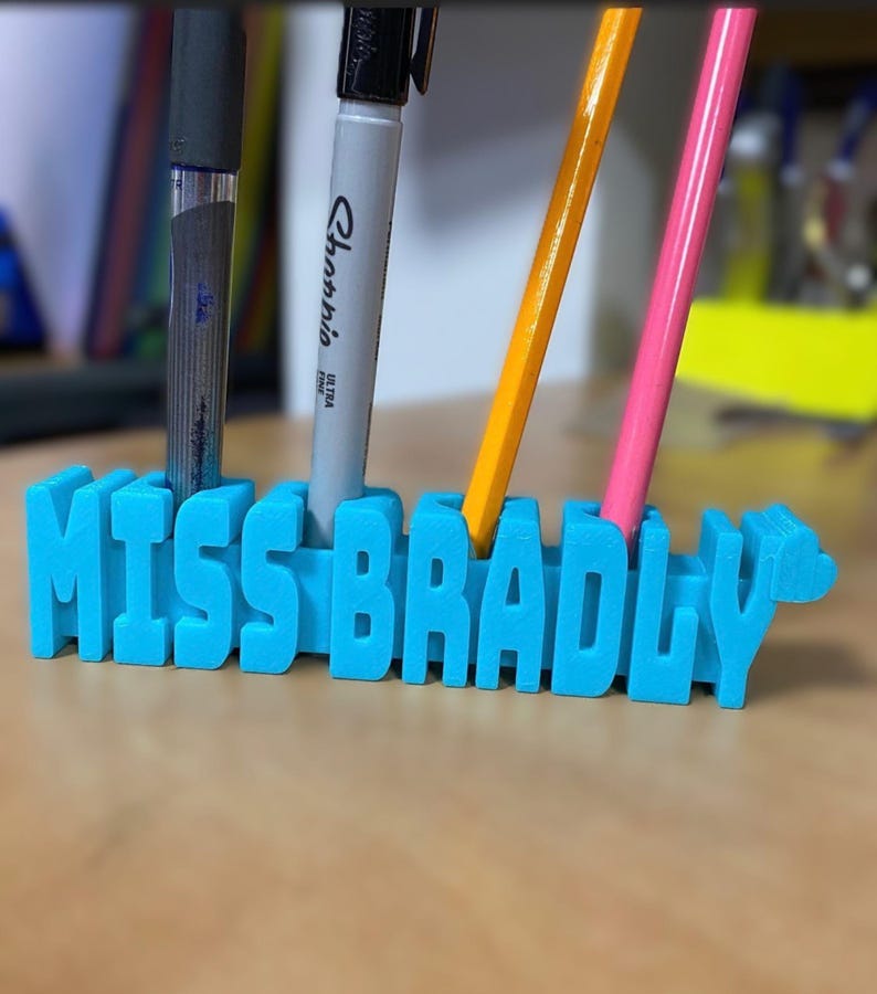 May include: A bright blue 3D printed desk organizer featuring the name MISS BRADLY in raised block letters. The organizer has four vertical slots holding a black pen, a gray marker, an orange pencil, and a pink pencil in an upright position.