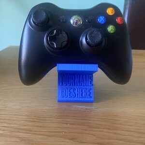 Può includere: Controller Xbox 360 nero appoggiato su un supporto blu. Il supporto presenta la scritta "YOURNAME GOES HERE". Il controller ha pulsanti colorati e due levette analogiche.