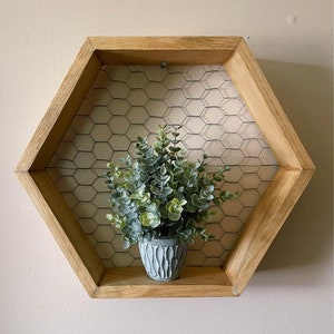 May include: A hexagonal wooden shelf with a wire mesh back, featuring a small potted plant. The shelf is stained a light brown colour and has a rustic, farmhouse style.