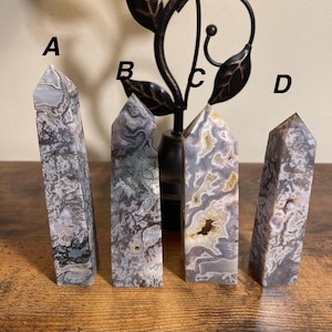May include: Four polished crystal obelisks with varying patterns and colors. The crystals are primarily shades of gray, white, and brown, with unique veining and textures. Each obelisk has a pointed top and a rectangular base, displayed on a wooden surface.