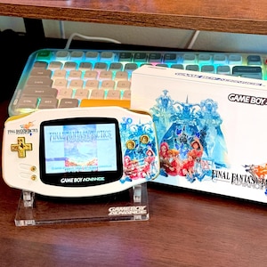 May include: A white Game Boy Advance handheld console with gold buttons and a Final Fantasy Tactics Advance screen display, positioned on a clear stand next to its original white retail box featuring character artwork and the game title.