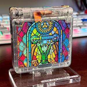 May include: A clear Game Boy Advance SP with a stained-glass design featuring a character holding a sword. The design includes vibrant colors like red, yellow, blue, and green. The base reads "GAME BOY ADVANCE SP".