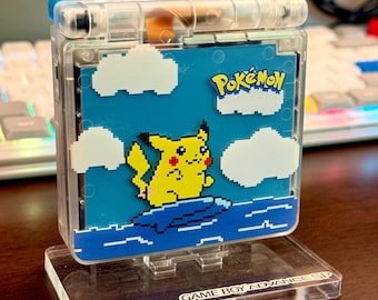 May include: A clear, transparent Game Boy Advance SP handheld console featuring a custom faceplate with a pixelated yellow Pikachu surfing on a blue wave against a blue sky with white clouds and the Pokémon logo, resting on a clear acrylic stand.