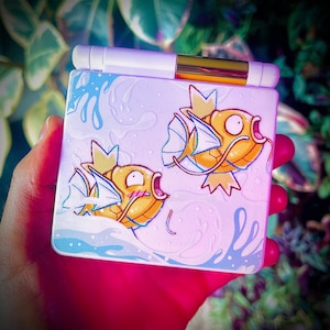 May include: A white and gold compact case featuring two cartoon goldfish with orange and yellow scales, blue fins, and a water wave design. The case has a white top with a gold accent.