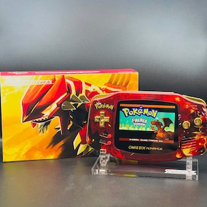 May include: A red and gold Game Boy Advance SP with a fiery design. The game cartridge is labeled "Pokémon FireRed Version".