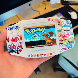 May include: A white Game Boy Advance SP with a custom Pokemon FireRed Version skin. The screen shows the game's title screen with the text "Pokemon FireRed Version" and the copyright information "© 2004 GAME FREAK inc.". The console has a purple and white button and a white D-pad. The skin features various Pokemon characters, including Charizard, Bulbasaur, Squirtle, and Pikachu.