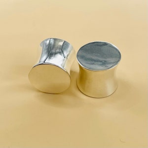 May include: Two silver ear gauges, one is slightly larger than the other. The gauges are smooth and have a cylindrical shape.