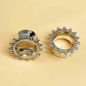 Puede incluir: Un par de dilatadores de oreja plateados con un diseño de túnel circular y un borde exterior decorativo adornado con pequeñas cuentas esféricas espaciadas uniformemente que crean una apariencia de sol alrededor de cada pieza de joyería.