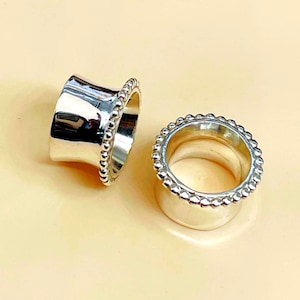 Peut inclure: Deux écarteurs d'oreille de couleur argentée avec un corps cylindrique concave lisse et un bord perlé décoratif le long du rebord extérieur. La surface métallique est polie et réfléchissante, offrant une finition brillante et uniforme.