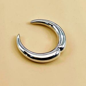 May include: A polished silver-tone ring with an open, crescent moon-shaped design. The band features a smooth, reflective metallic surface that tapers at both ends, creating a continuous, curved silhouette that remains open at the top.