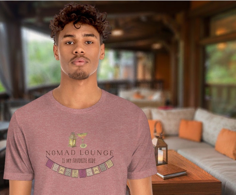 K&ouml;nnte beinhalten: Ein Mann tr&auml;gt ein heidefarbenes T-Shirt mit dem Text "Nomad Lounge is my favorite ride" und einer Grafik einer Laterne und eines Cocktailglases mit einer Girlande aus bunten Flaggen.