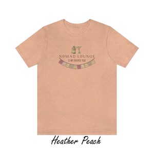 K&ouml;nnte beinhalten: Ein T-Shirt in der Farbe Heidelbeer-Pfirsich mit einem grafischen Design aus einer Laterne, einem Margarita-Glas und einer Girlande aus bunten Flaggen. Der Text "Nomad Lounge is my favorite ride" ist auf das T-Shirt gedruckt.