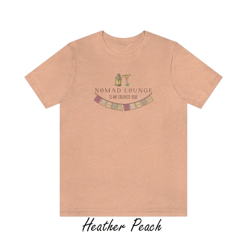 K&ouml;nnte beinhalten: Ein T-Shirt in der Farbe Heidelbeer-Pfirsich mit einem grafischen Design aus einer Laterne, einem Margarita-Glas und einer Girlande aus bunten Flaggen. Der Text "Nomad Lounge is my favorite ride" ist auf das T-Shirt gedruckt.