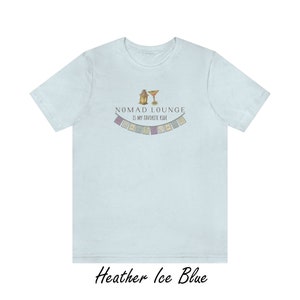 K&ouml;nnte beinhalten: Ein hellblaues T-Shirt mit einem Aufdruck einer Laterne und eines Cocktailglases mit dem Text "Nomad Lounge is my favorite ride" und einer Reihe bunter Flaggen.