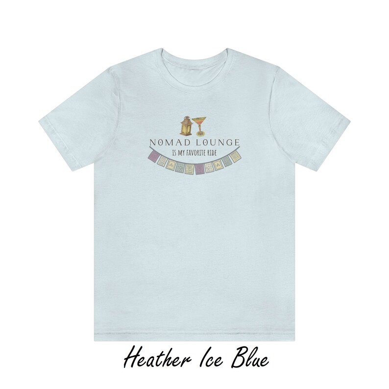 K&ouml;nnte beinhalten: Ein hellblaues T-Shirt mit einem Aufdruck einer Laterne und eines Cocktailglases mit dem Text "Nomad Lounge is my favorite ride" und einer Reihe bunter Flaggen.