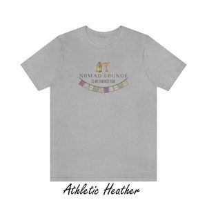 K&ouml;nnte beinhalten: Ein heidefarbenes T-Shirt mit dem Text "Nomad Lounge is my favorite ride" und einer Grafik einer Laterne und eines Margarita-Glases mit einer Girlande aus bunten Flaggen &uuml;ber dem Text.