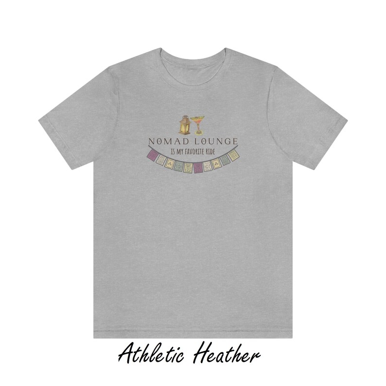 K&ouml;nnte beinhalten: Ein heidefarbenes T-Shirt mit dem Text "Nomad Lounge is my favorite ride" und einer Grafik einer Laterne und eines Margarita-Glases mit einer Girlande aus bunten Flaggen &uuml;ber dem Text.