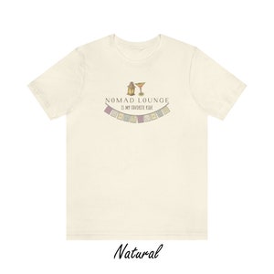 K&ouml;nnte beinhalten: Ein beiges T-Shirt mit einem grafischen Design einer Laterne und eines Cocktailglases mit dem Text "Nomad Lounge is my favorite ride" und einer Reihe bunter Flaggen.