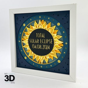 Può includere: Un'opera d'arte in carta a strati incorniciata, raffigurante un'eclissi solare totale. L'opera ha una cornice bianca e uno sfondo blu scuro con stelle. Il sole è raffigurato in tonalità di giallo e oro, con il testo "TOTAL SOLAR ECLIPSE 04.08.2024" al centro.