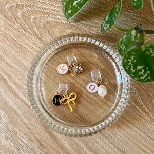May include: Three gold phone charms with different designs. The first charm has a white circle with the letter "M" in gold. The second charm has a black circle with the letter "N" in gold and a gold bow. The third charm has a white circle with the letter "O" in gold and a white pearl.
