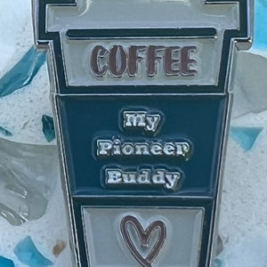 May include: A silver metal coffee cup pin with a white and blue design. The cup says "COFFEE" in pink letters and "My Pioneer Buddy" in white letters. The bottom of the cup has a pink heart.