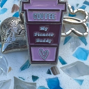 May include: A silver metal keychain with a pink and brown coffee cup charm. The charm has the word "COFFEE" printed on it and the words "My Pioneer Buddy" printed below it. The charm also has a pink heart on the bottom.