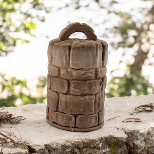 May include: A brown, cylindrical decorative object resembling a miniature well. The object is made of a material that looks like stone, with a handle at the top. The well is sitting on a stone surface.