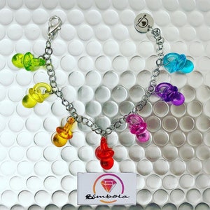 May include: A silver chain bracelet with colorful plastic pacifier charms. The charms are in the colors of the rainbow, with a yellow, orange, red, purple, blue, and green pacifier. The bracelet has a silver clasp and a small silver charm with a diamond shape.