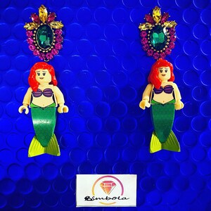 May include: A pair of earrings featuring a mermaid figurine with a green tail and a crown of jewels. The mermaid is wearing a purple seashell bra. The earrings are on a blue background.