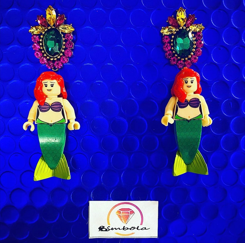 May include: A pair of earrings featuring a mermaid figurine with a green tail and a crown of jewels. The mermaid is wearing a purple seashell bra. The earrings are on a blue background.