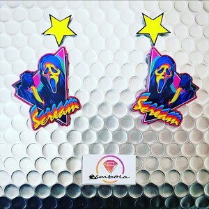 Könnte beinhalten: Zwei bunte Scream-Film-Ohrringe mit einem gelben Stern-Anhänger. Die Ohrringe zeigen ein Schwarzweißbild von Ghostface mit einem Messer und das Wort "Scream" in einer Regenbogen-Schrift.