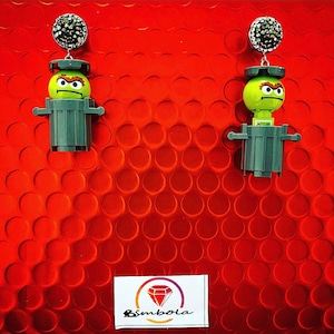 Puede incluir: Un par de pendientes verdes y grises con un personaje de dibujos animados con una expresión de enfado. El personaje lleva una papelera en la cabeza. Los pendientes están adornados con detalles plateados brillantes.