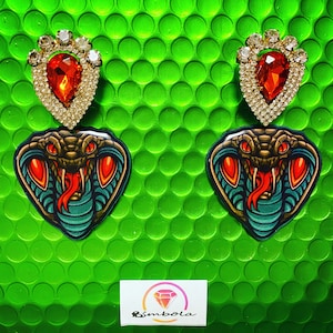 Könnte beinhalten: Ein Paar Hängeohrringe mit einem roten Strassstein und einem grünen Schlangenmotiv. Die Ohrringe befinden sich auf einem grünen Hintergrund mit einem weißen Logo, auf dem "Bimboia" steht.