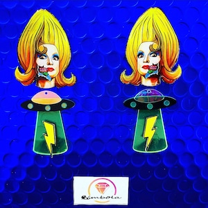 Puede incluir: Dos pares de pendientes con una mujer de dibujos animados con cabello amarillo y una nave espacial extraterrestre verde. La cara de la mujer está parcialmente oscurecida por una criatura extraterrestre verde. Los pendientes están sobre un fondo azul.